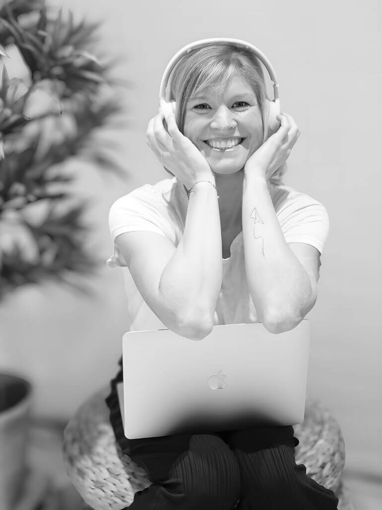 Sunshine Podcast Produktion Beratung Sunshine VA mit weißen Kopfhörern, die sie sich rechts und links festhält. Sitzt auf einem Korb-Puff und lehnt sich an ihrem Laptop an